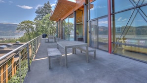 A balcony with tables and chairs, large glass windows, and a view of the landscape. - Alizes - Luxury, Flagship Best Gorge Views, Downtown, Theater Room, Exceptional (Hood River)