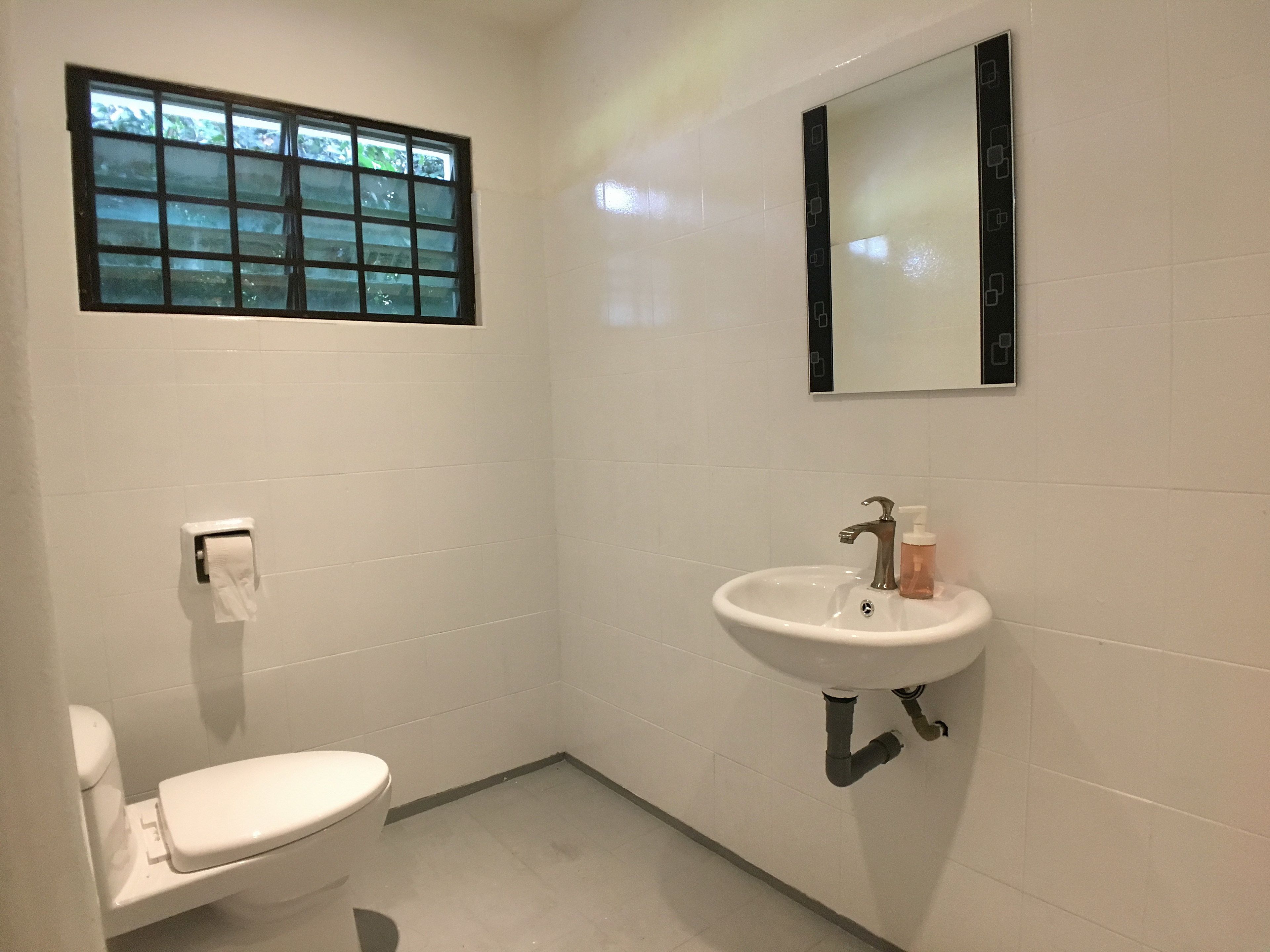 A small, clean bathroom with white tiles, a toilet, a sink with a mirror above it, and a window with frosted glass.