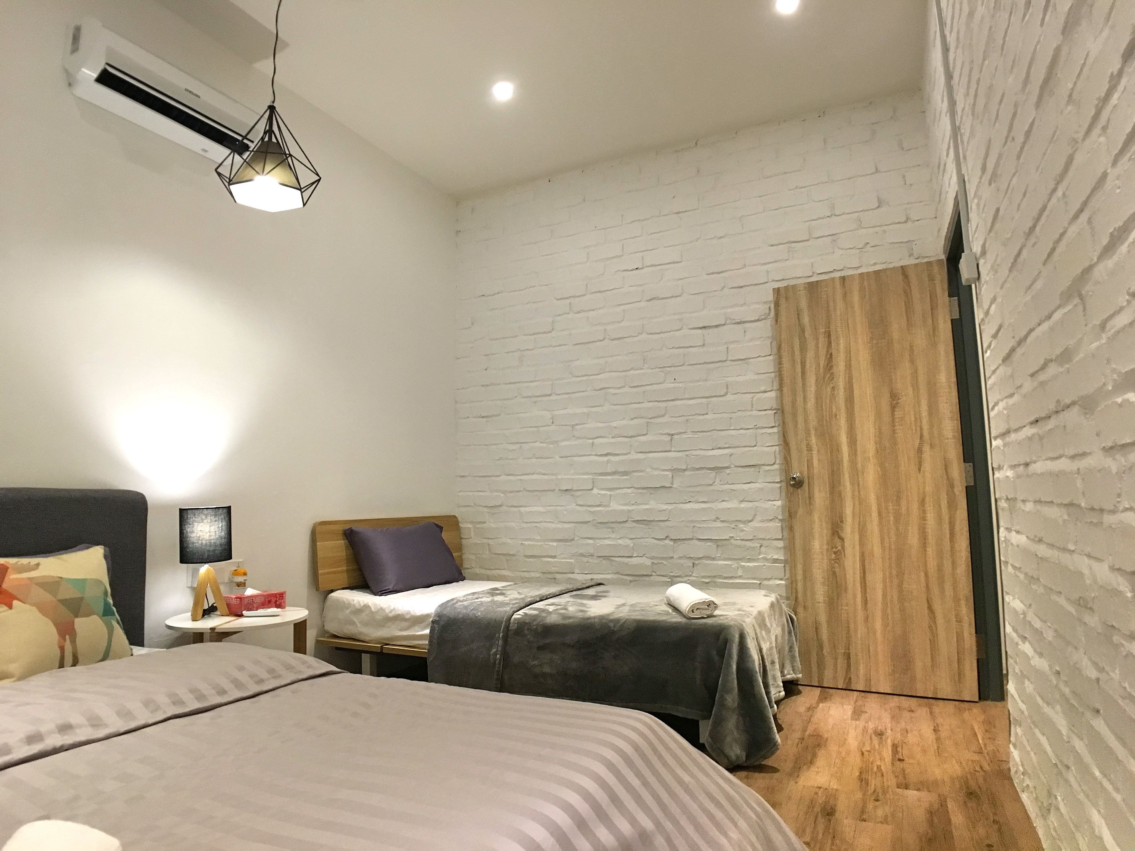 A bedroom with a bed, a bedside table, a lamp, a wall-mounted air conditioner, and a wooden door.