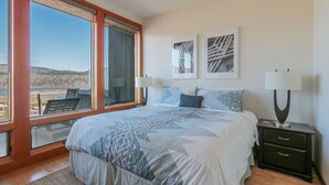 Un dormitorio con una cama grande, dos mesitas de noche con lámparas, vistas al exterior a través de un ventanal amplio y dos cuadros enmarcados en la pared.