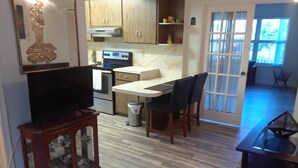 Cocina compacta con estufa, horno y armarios de madera. Cuenta con un comedor con mesa y sillas, y un televisor sobre un mueble.