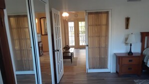 Una habitación con piso de madera, una cama con cabecera, una mesita de noche con lámpara y una puerta que da a otra habitación.