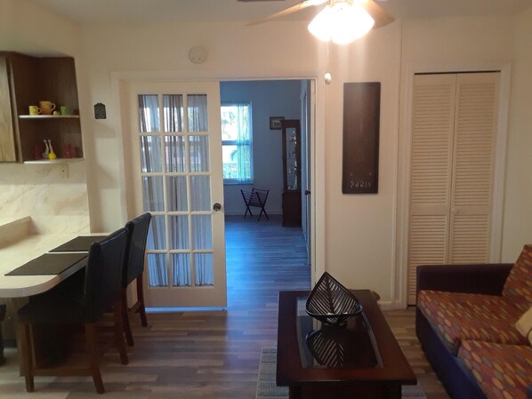 A living room with a sofa, a coffee table, and a dining area with a table and chairs. There is a door leading to another room.