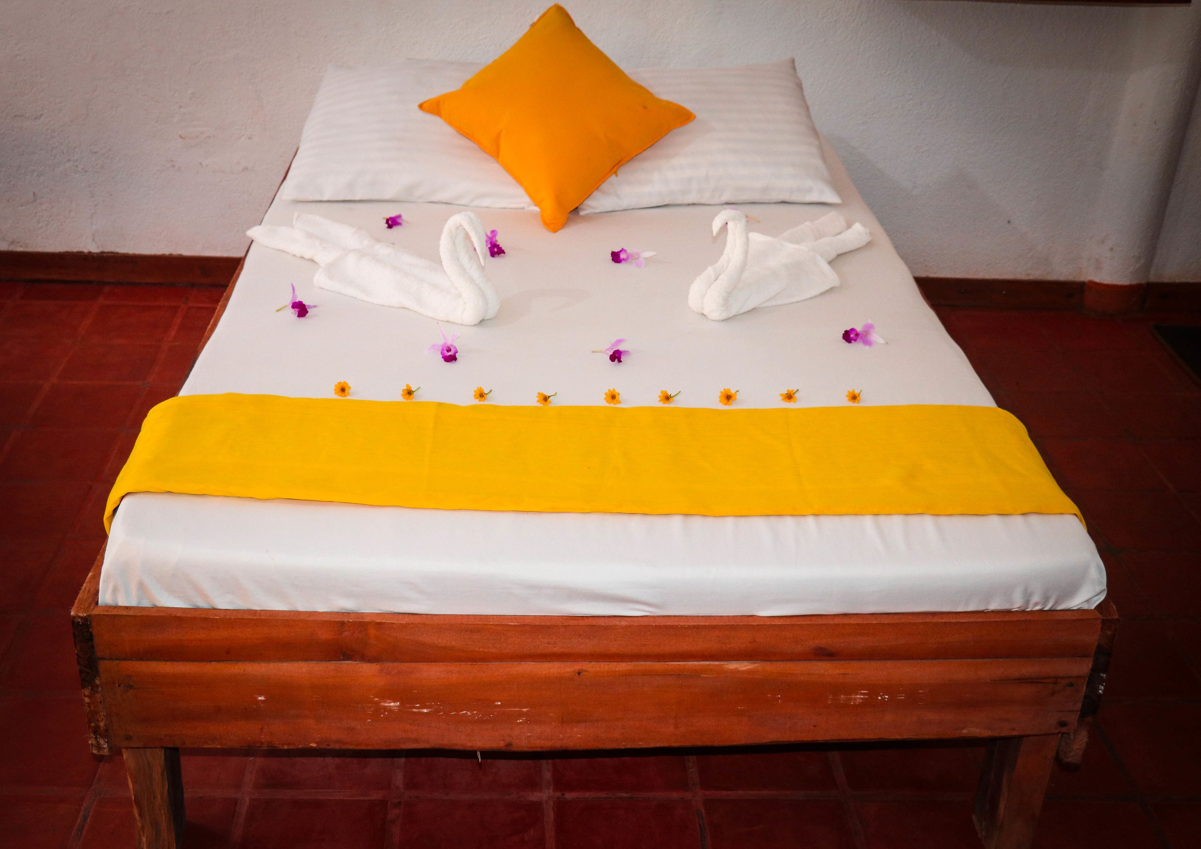 Een bed met een witte lakenset, een oranje kussen, twee witte handdoeken in de vorm van zwanen en een gele deken.