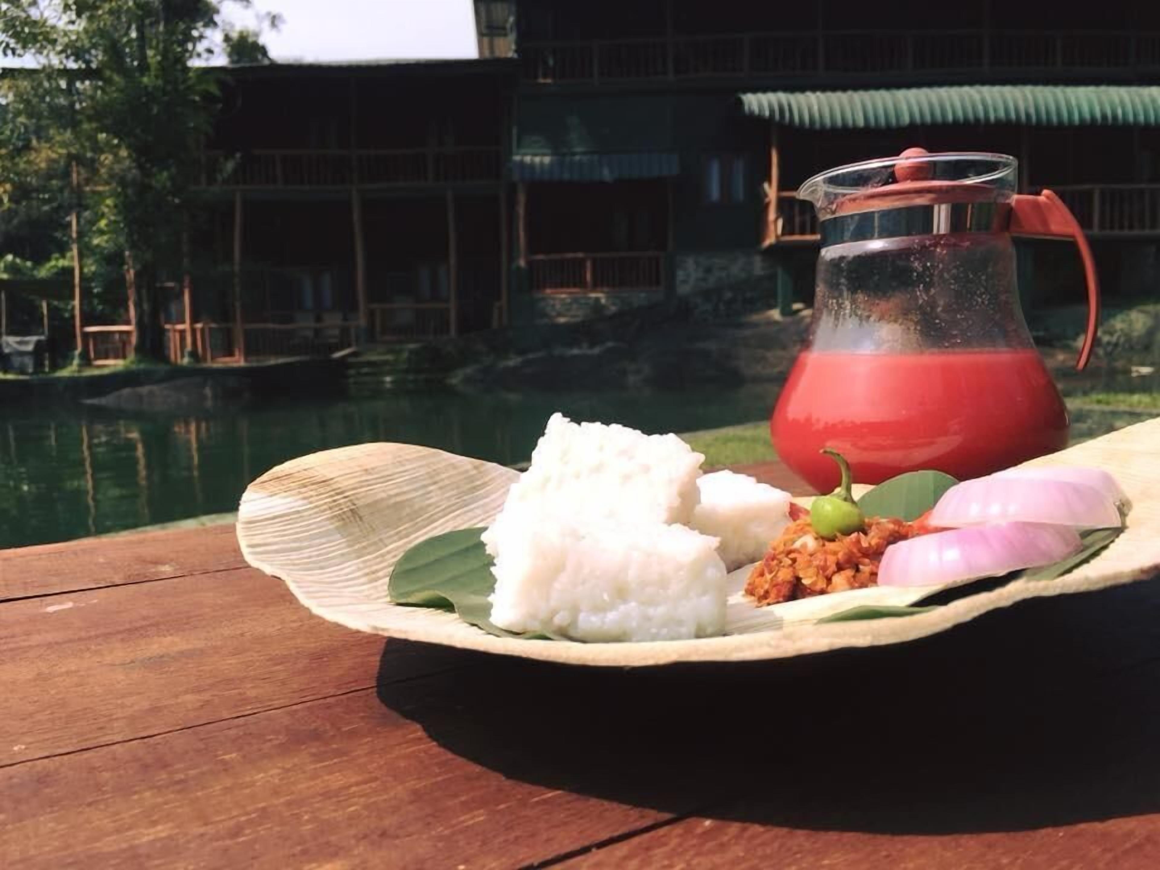 A wooden table with a plate of food, including rice, a green chili, and a red drink in a glass pitcher.
