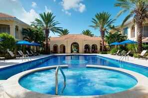 A swimming pool with lounge chairs and umbrellas, surrounded by palm trees and a building with a tiled roof.