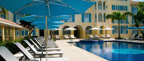 Ein Poolbereich mit Liegestühlen und Sonnenschirmen, ein Schwimmbecken und im Hintergrund ein großes Gebäude mit blauen Fensterläden.