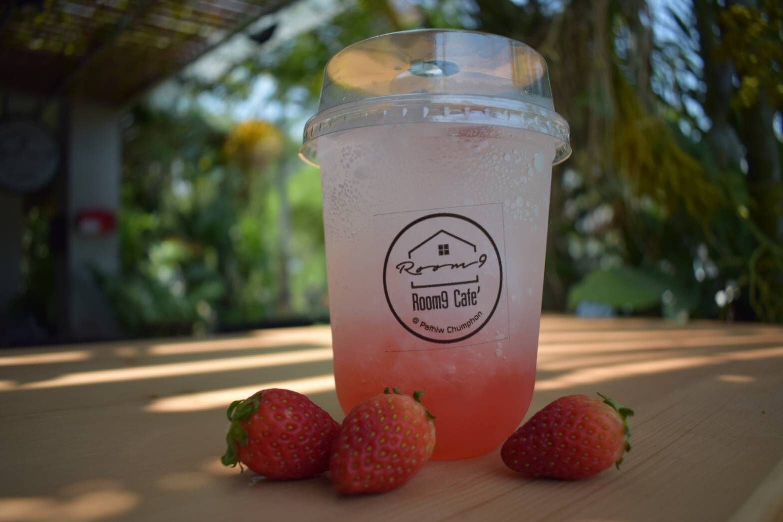 Ein durchsichtiges Plastikbecher mit einem Aufkleber mit der Aufschrift 'Room9 Cafe' und einem Logo, platziert auf einer Holzoberfläche mit drei Erdbeeren daneben.
