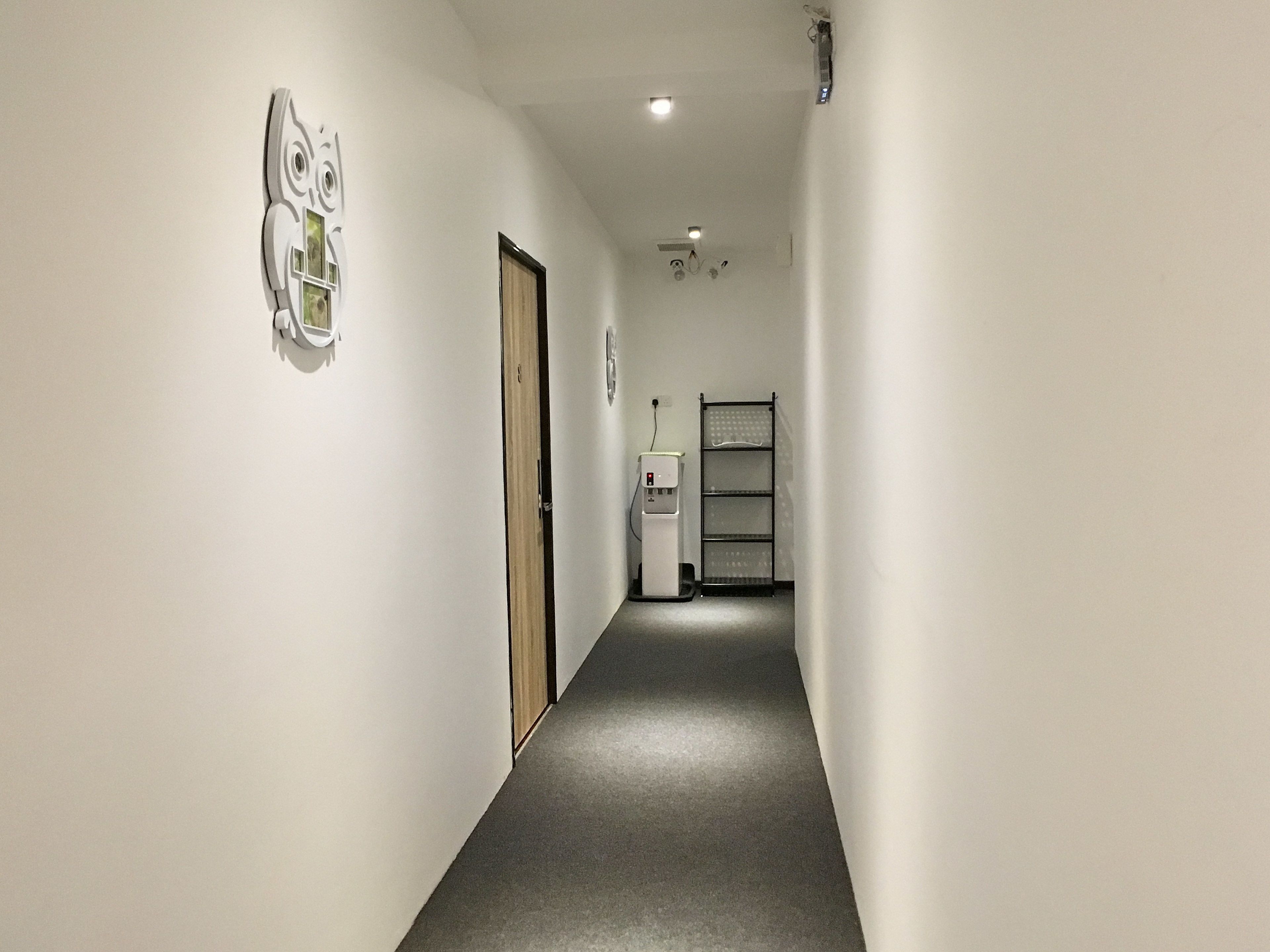 Un couloir étroit, recouvert d’un tapis gris, avec une porte sur la gauche et un distributeur d’eau sur la droite.
