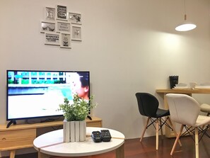 A modern living room with a flat-screen TV, a round coffee table with a plant and remotes, two chairs, and a dining area with a table and chairs. - iBook7 - 2 Bedroom Gurney Studio Suite by iBook Homestay (George Town)