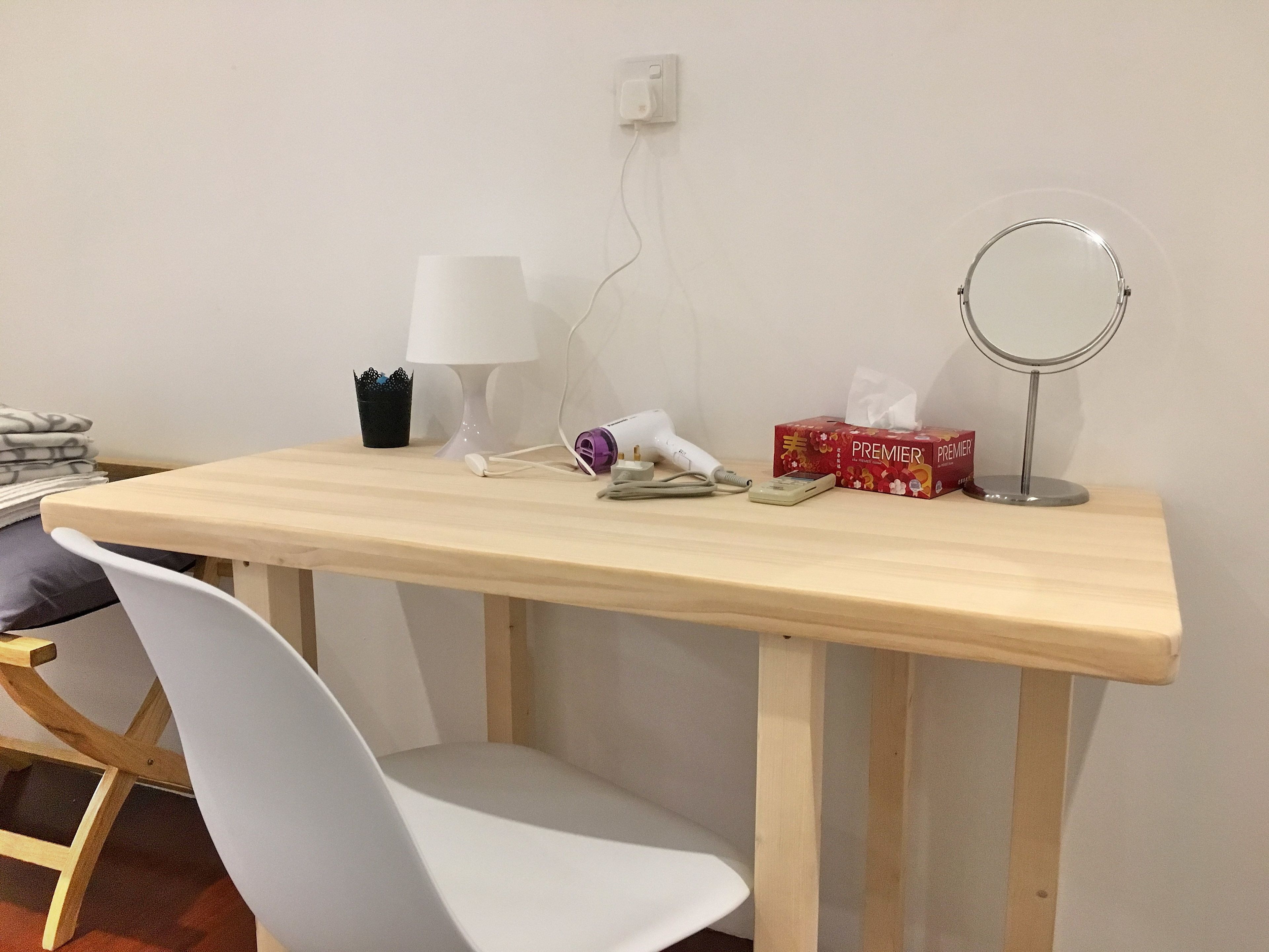 A wooden desk with a white chair, a lamp, a hairdryer, a tissue box, and a compact mirror.