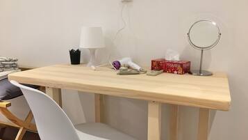 A wooden desk with a white chair, a lamp, a hairdryer, a tissue box, and a compact mirror.