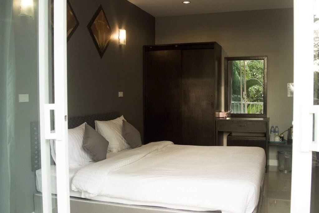 A bedroom with a bed, a wardrobe, a nightstand, and a window with a view of greenery.