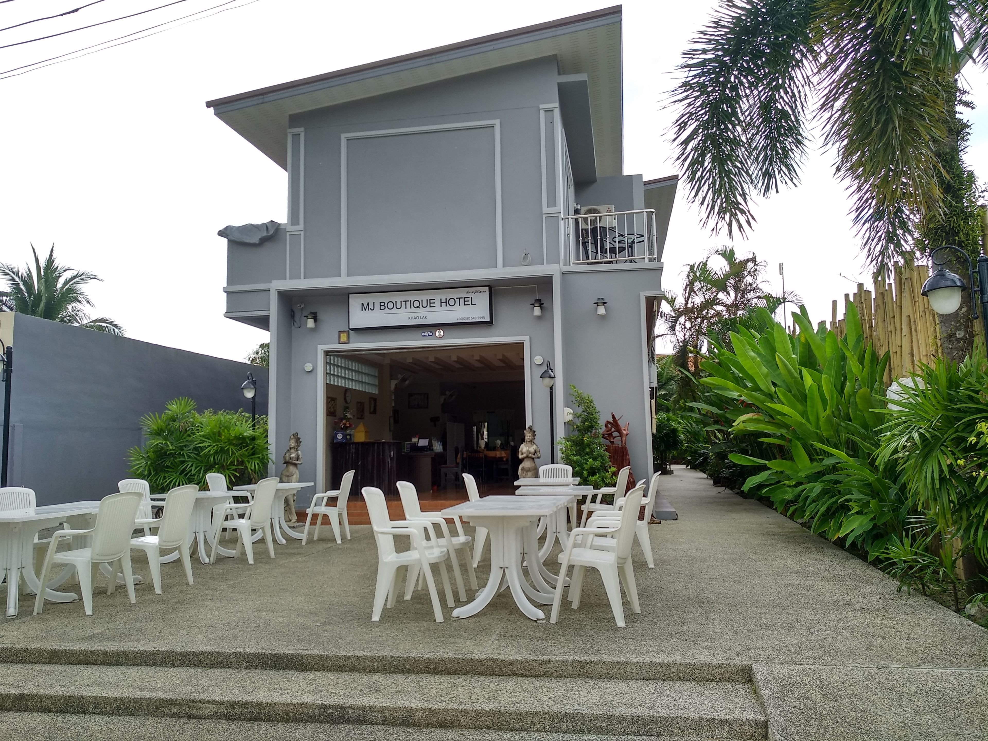 A two-story building with a sign reading "MJ Boutique Hotel." There are outdoor seating areas with white plastic chairs and tables.
