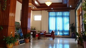 A spacious lobby with a wooden ceiling, a chandelier, and a large window with blue curtains. There are potted plants and a signboard visible. - Princess Hotel (Kengtung)