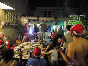 A group of people gathered around a table with food and drinks, wearing festive hats, in an outdoor setting at night. - The Oasis Hostel - Adults Only (Bangkok)