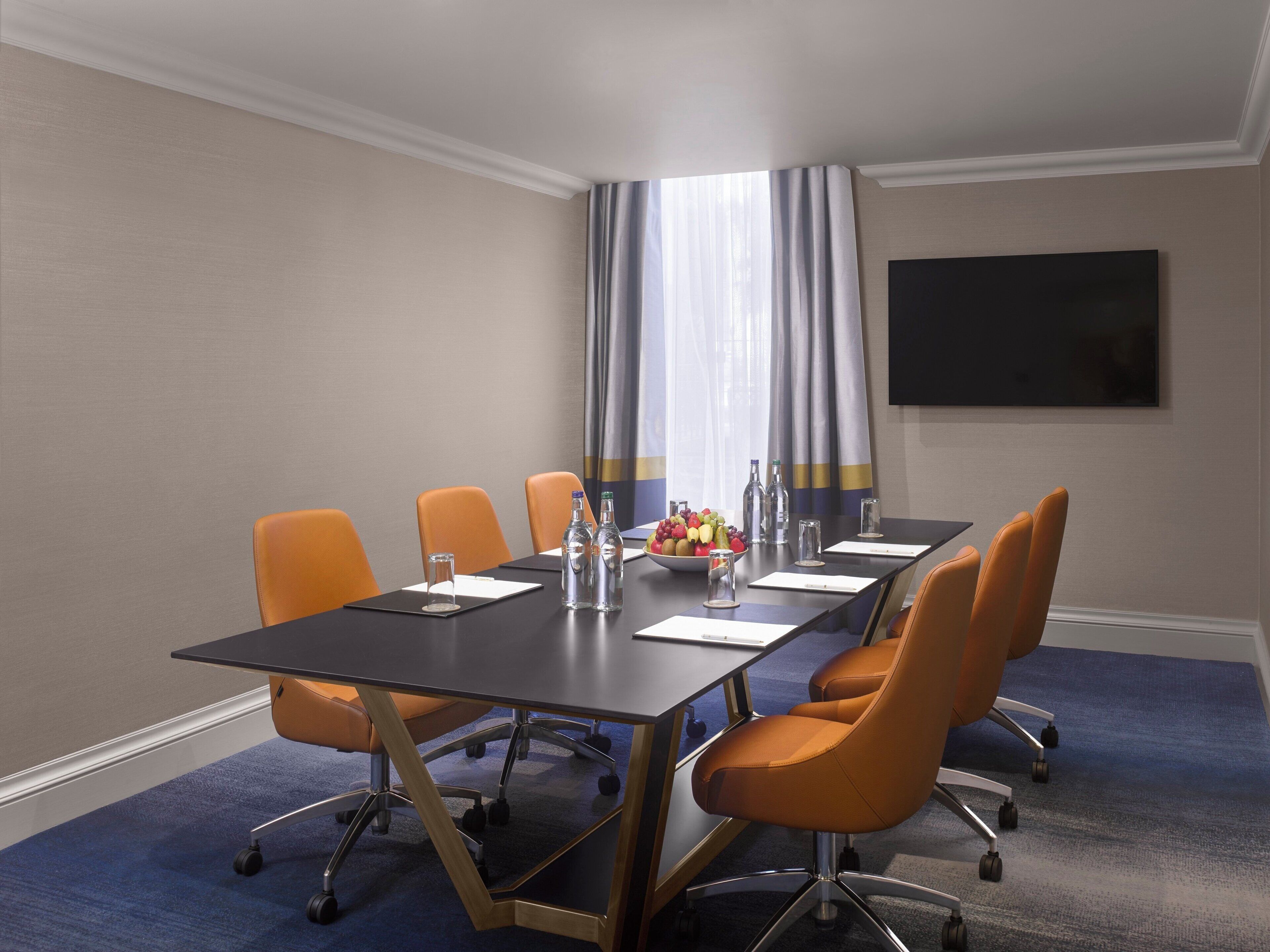 A conference room with a long table, six orange chairs, a fruit bowl, water bottles, and a flat-screen TV on the wall.
