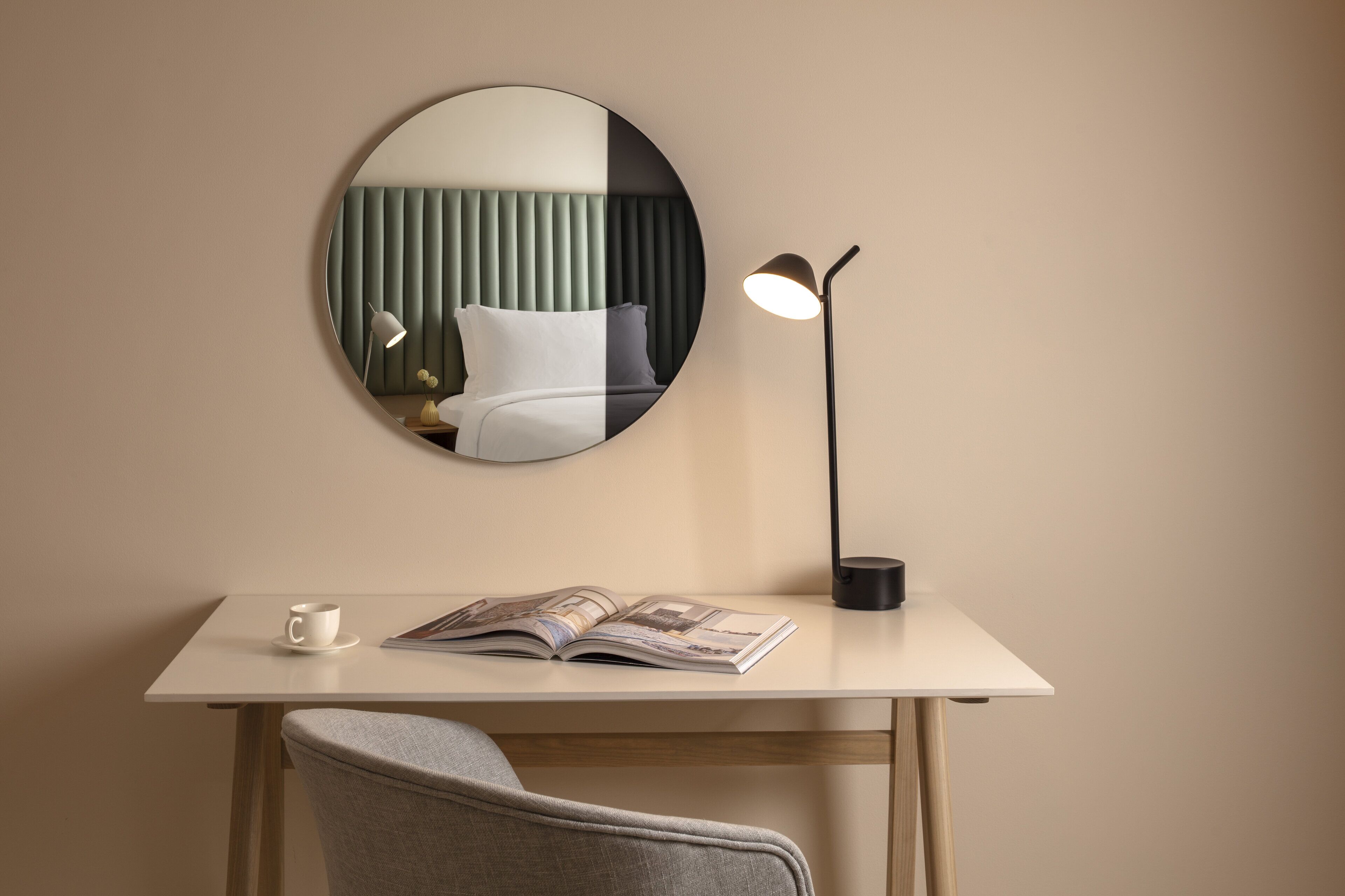 Una habitación moderna con un espejo redondo en la pared, una mesita de noche con lámpara, una silla y una revista.