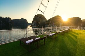 Um terraço com espreguiçadeiras e uma mesa, com vista para um corpo de água e montanhas ao fundo.