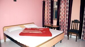 A single bed with a wooden frame and white bedding, a small wooden table beside it, and a chair with a dark cushion. - Hotel Golden (Haridwar)