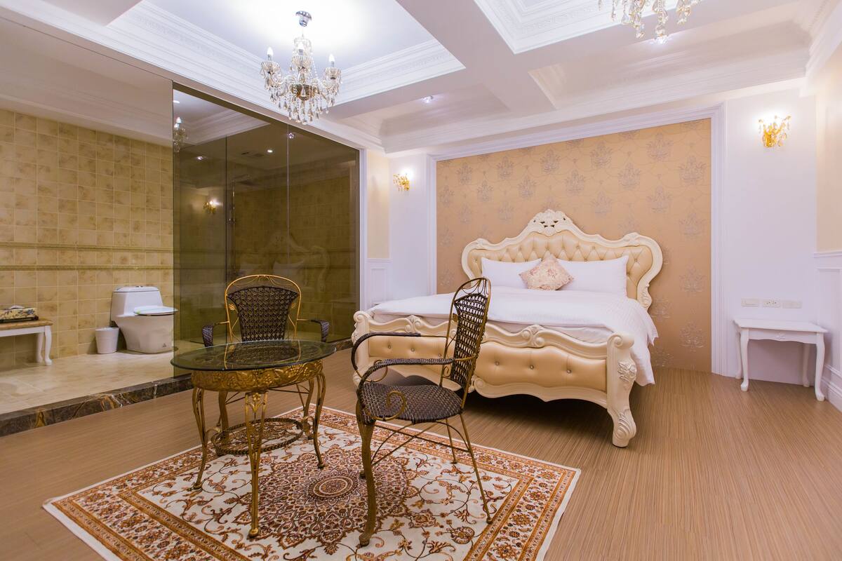A bedroom with a large bed, a glass shower, a dining table with chairs, and a chandelier.