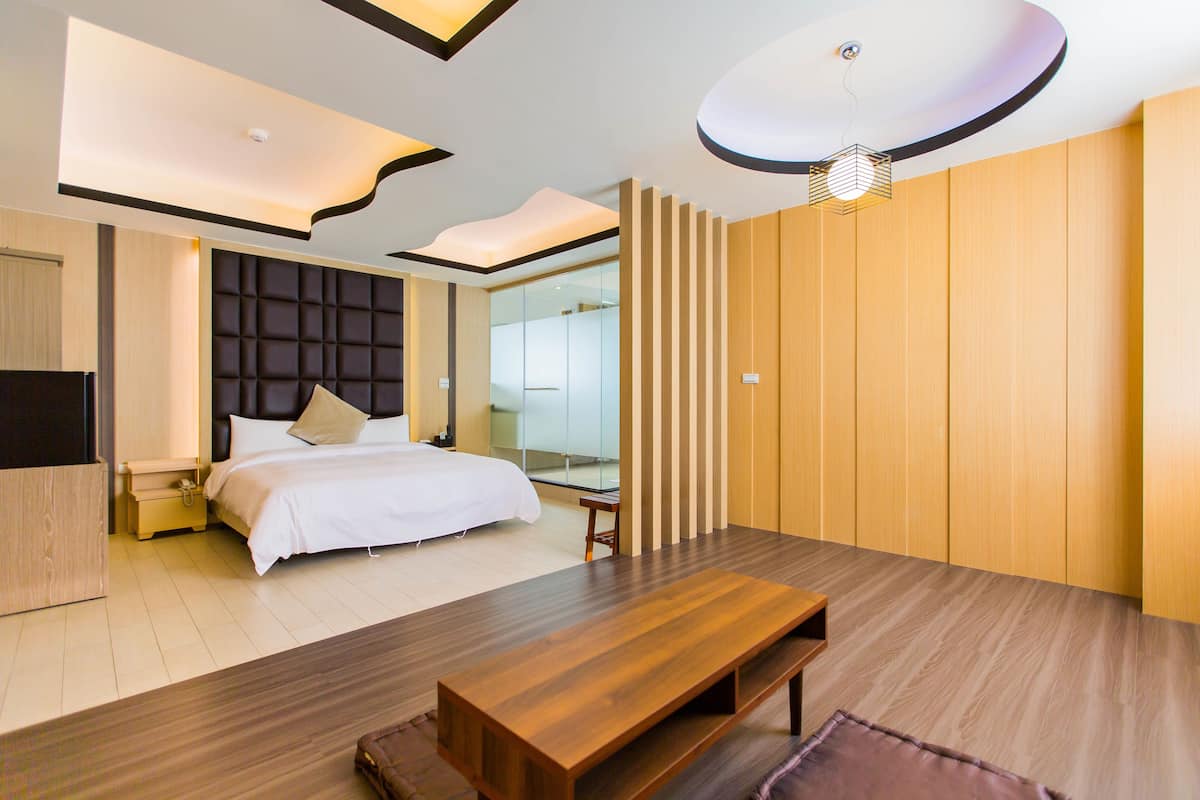 A modern hotel room with a large bed, wooden flooring, a built-in bench, a TV, and a unique ceiling design.