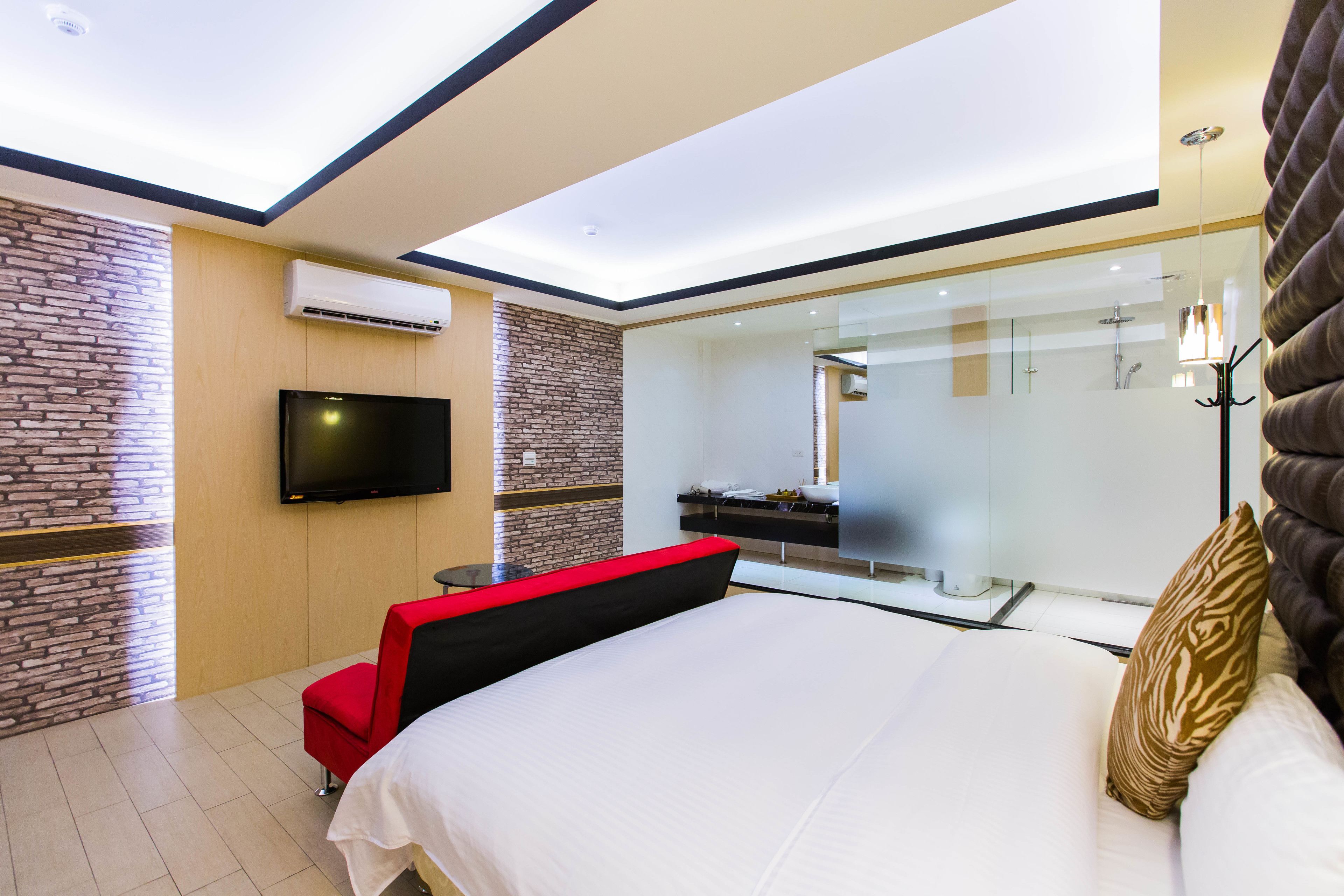 A modern hotel room with a large bed, a flat-screen TV, a glass shower enclosure, and a red chair.