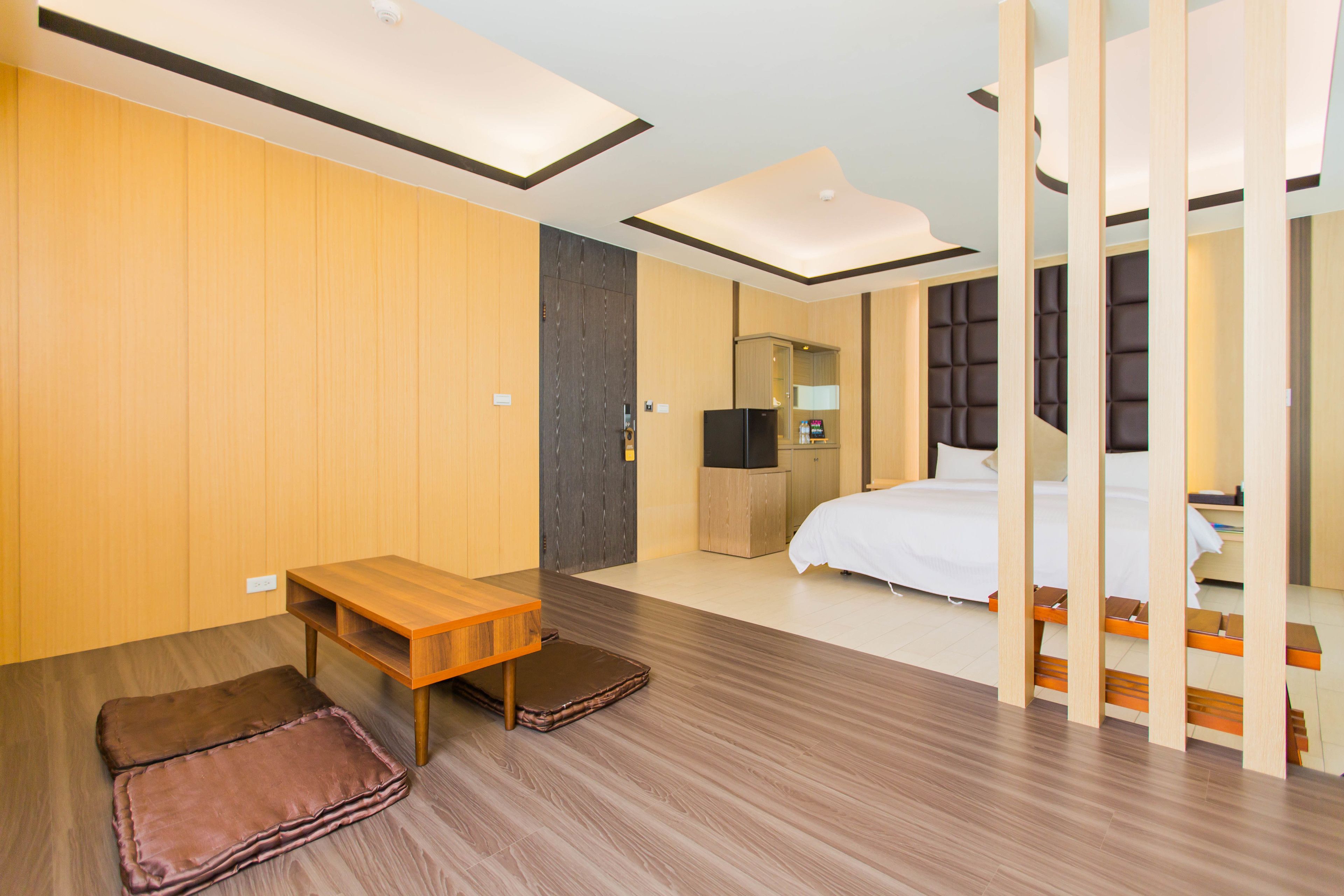 A modern hotel room with a wooden floor, a wooden desk, a bed with a white duvet, a wardrobe, and a small seating area with cushions.