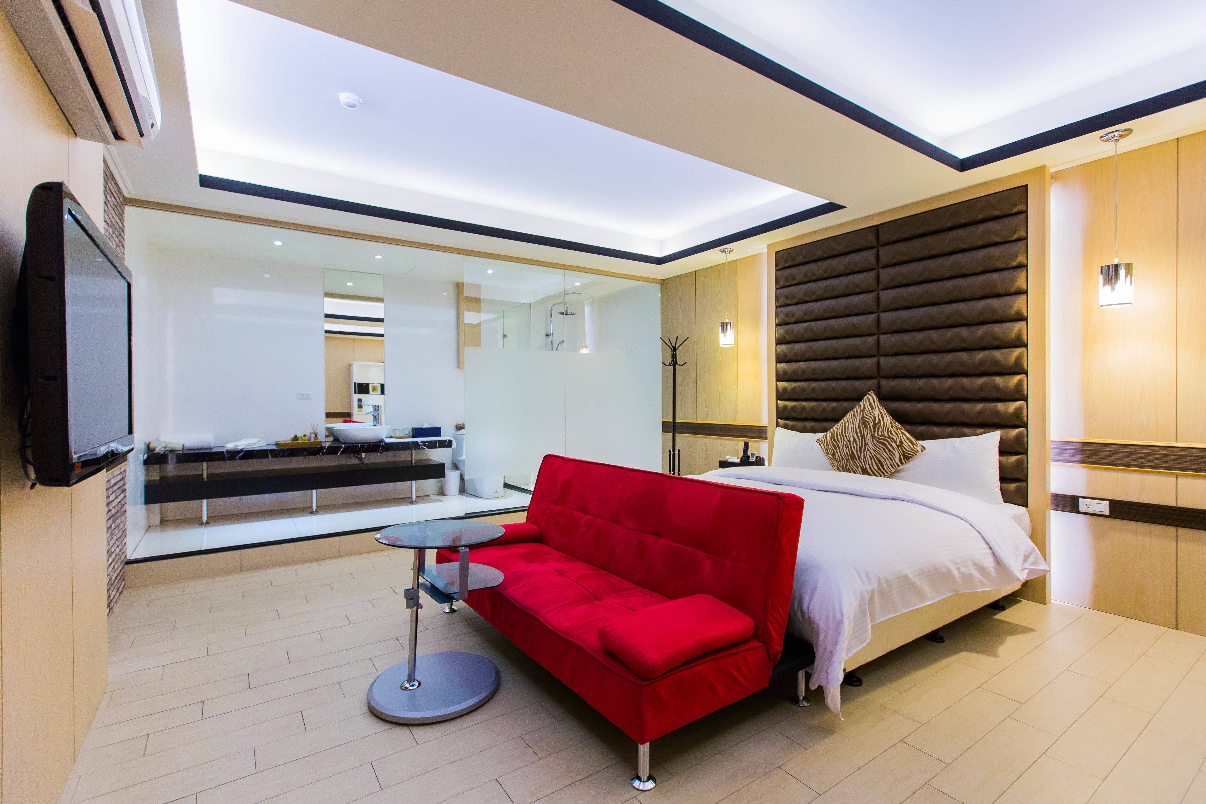 A modern hotel room with a red sofa, a bed with a wooden headboard, a large mirror, and a flat-screen TV mounted on the wall.