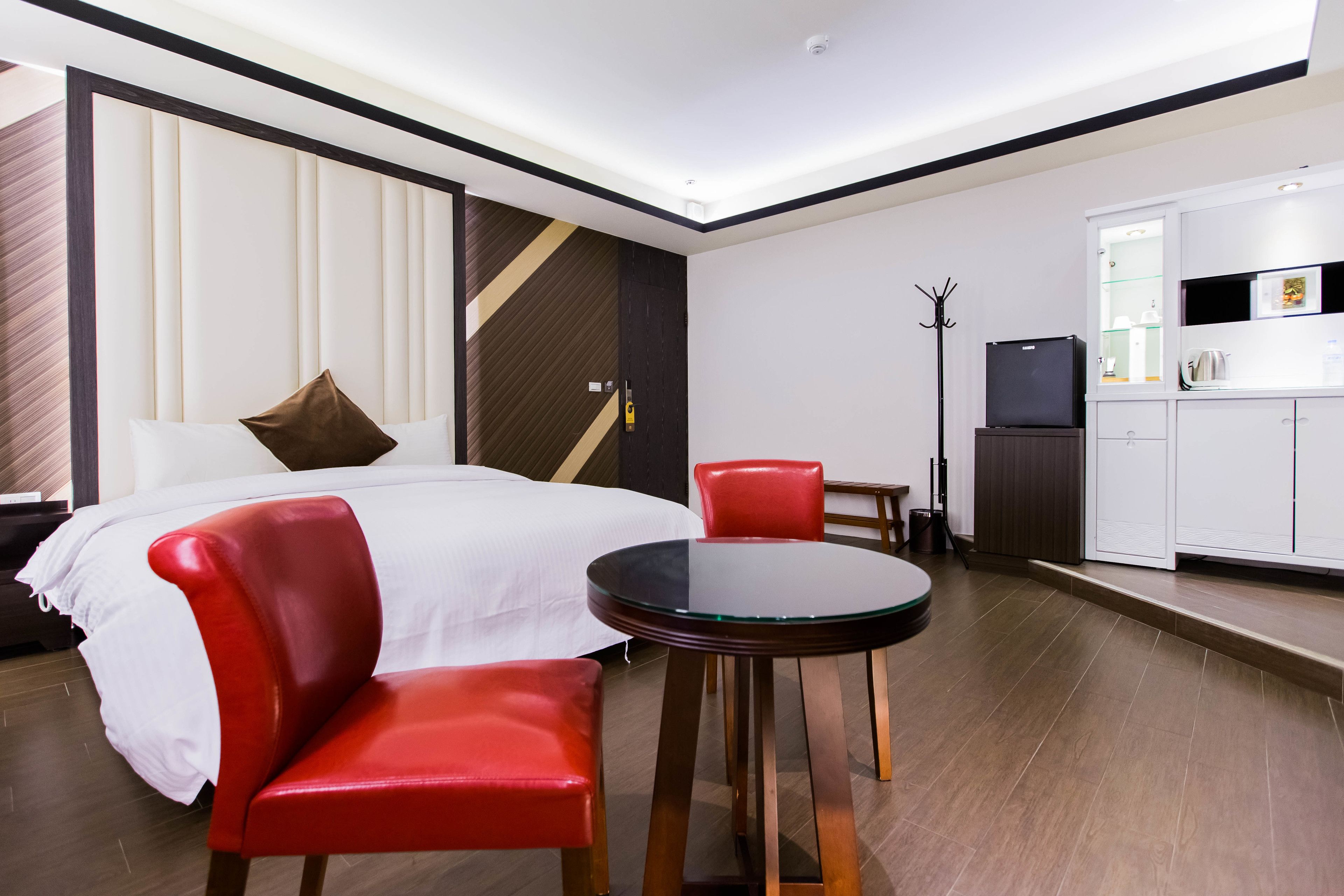 A hotel room with a bed, a round table with two red chairs, a small kitchenette, and a coat rack.