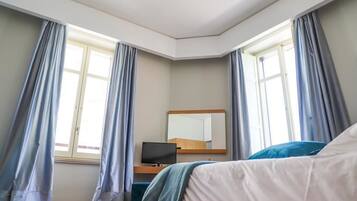 A bedroom with a bed, a desk with a computer, a mirror, and large windows with curtains.