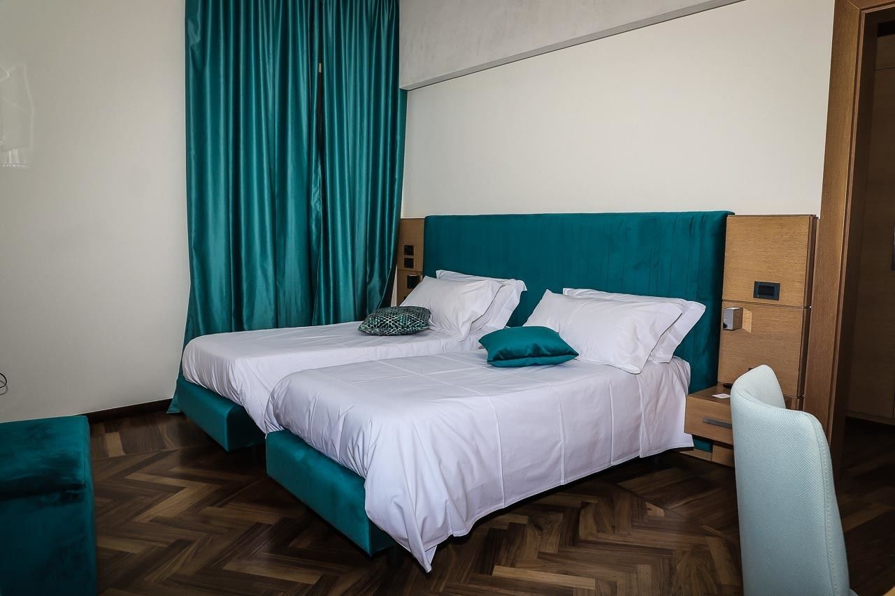 A double bed with teal bedding and pillows, a wooden headboard, a wooden nightstand, and a teal chair.