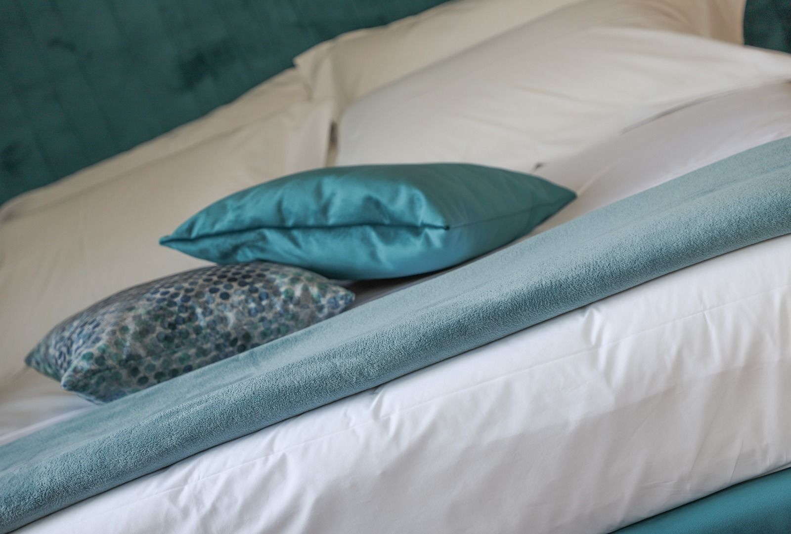 A bed with a teal bedspread, two pillows, and a patterned pillow.