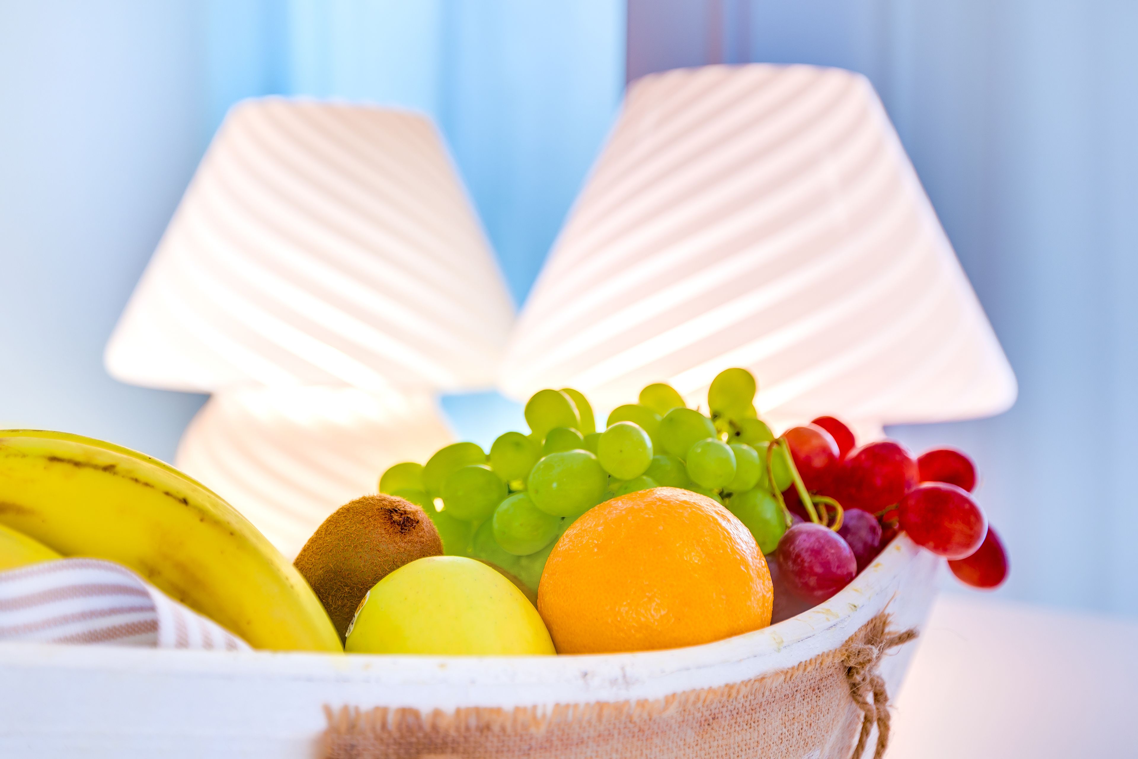 Un panier rempli de fruits, notamment des bananes, des raisins et une orange, placé devant une lampe au motif à rayures.