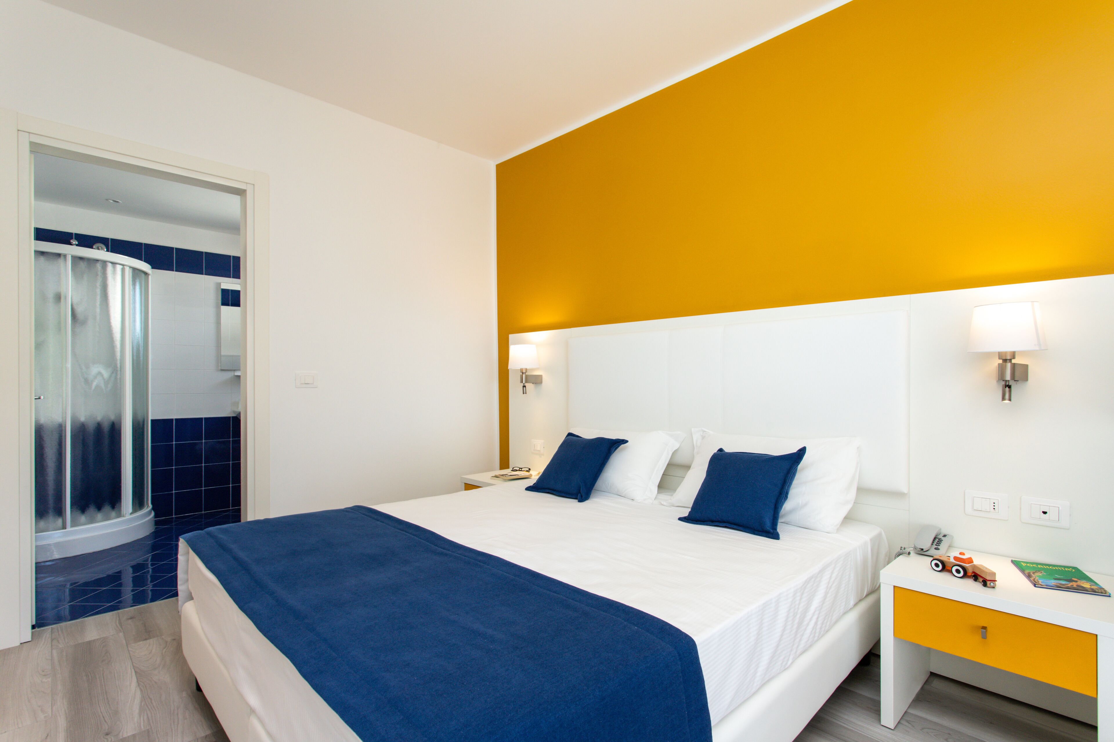 A hotel room with a bed, a nightstand, a bathroom with a shower, and a yellow accent wall.