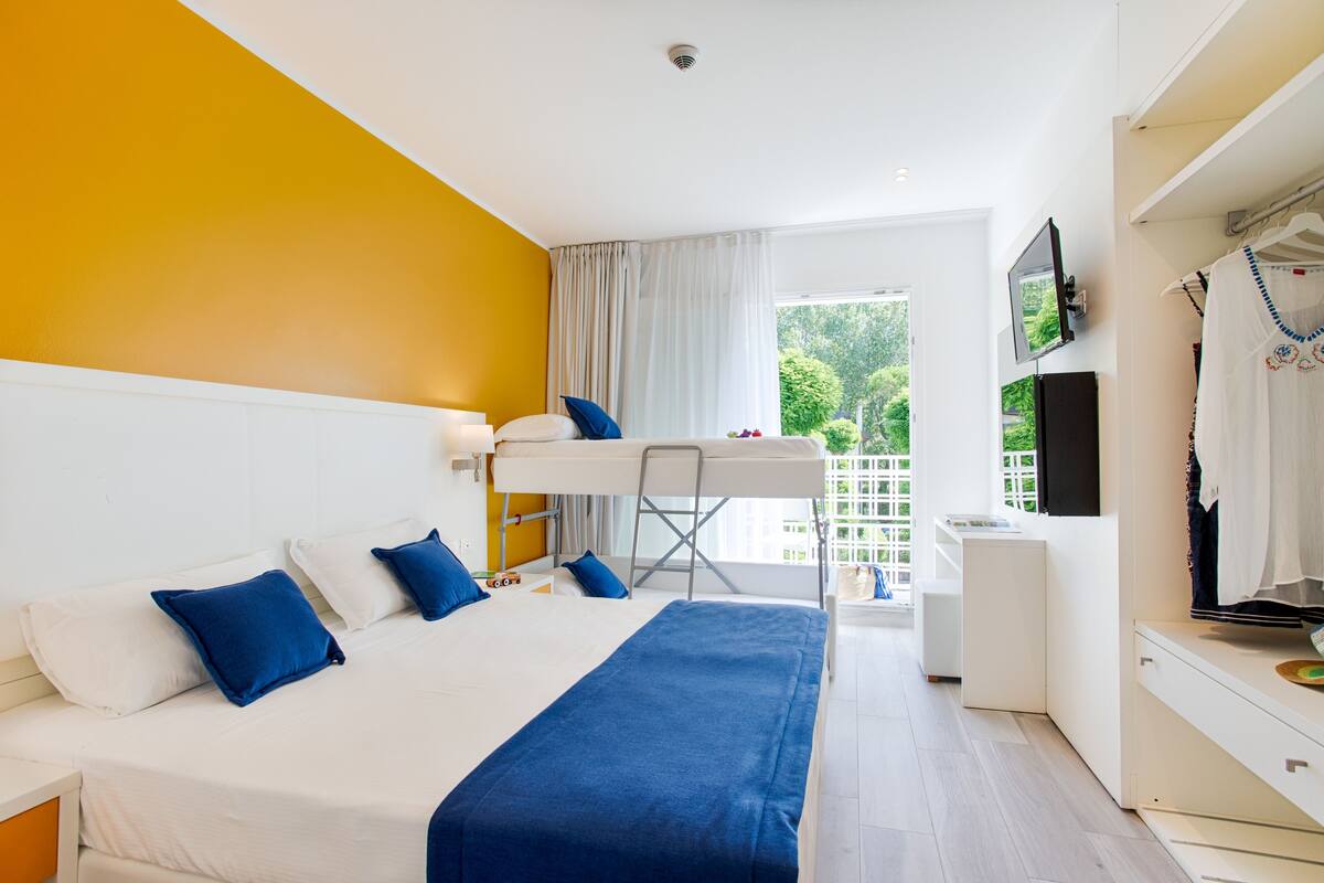 Habitación de hotel con literas, un escritorio, televisor y balcón con vistas a zonas verdes.
