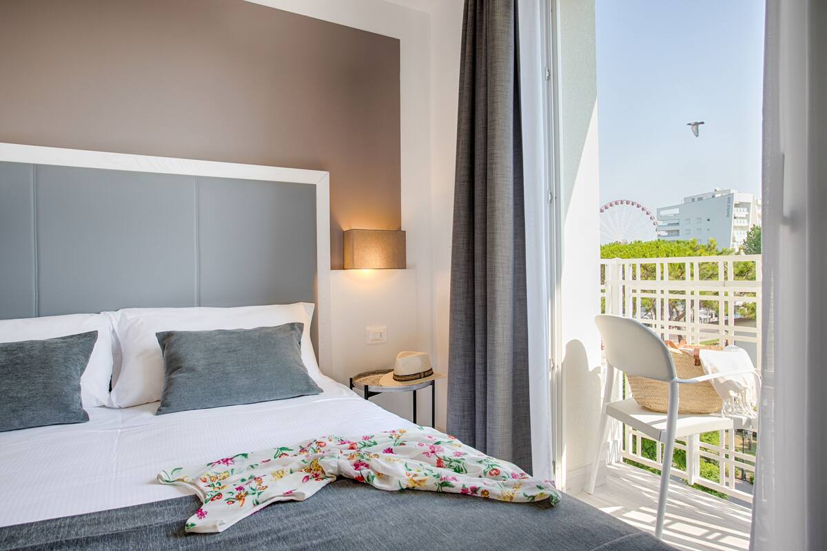 Una habitación de hotel con cama, mesita de noche, lámpara, silla y vista a una noria.