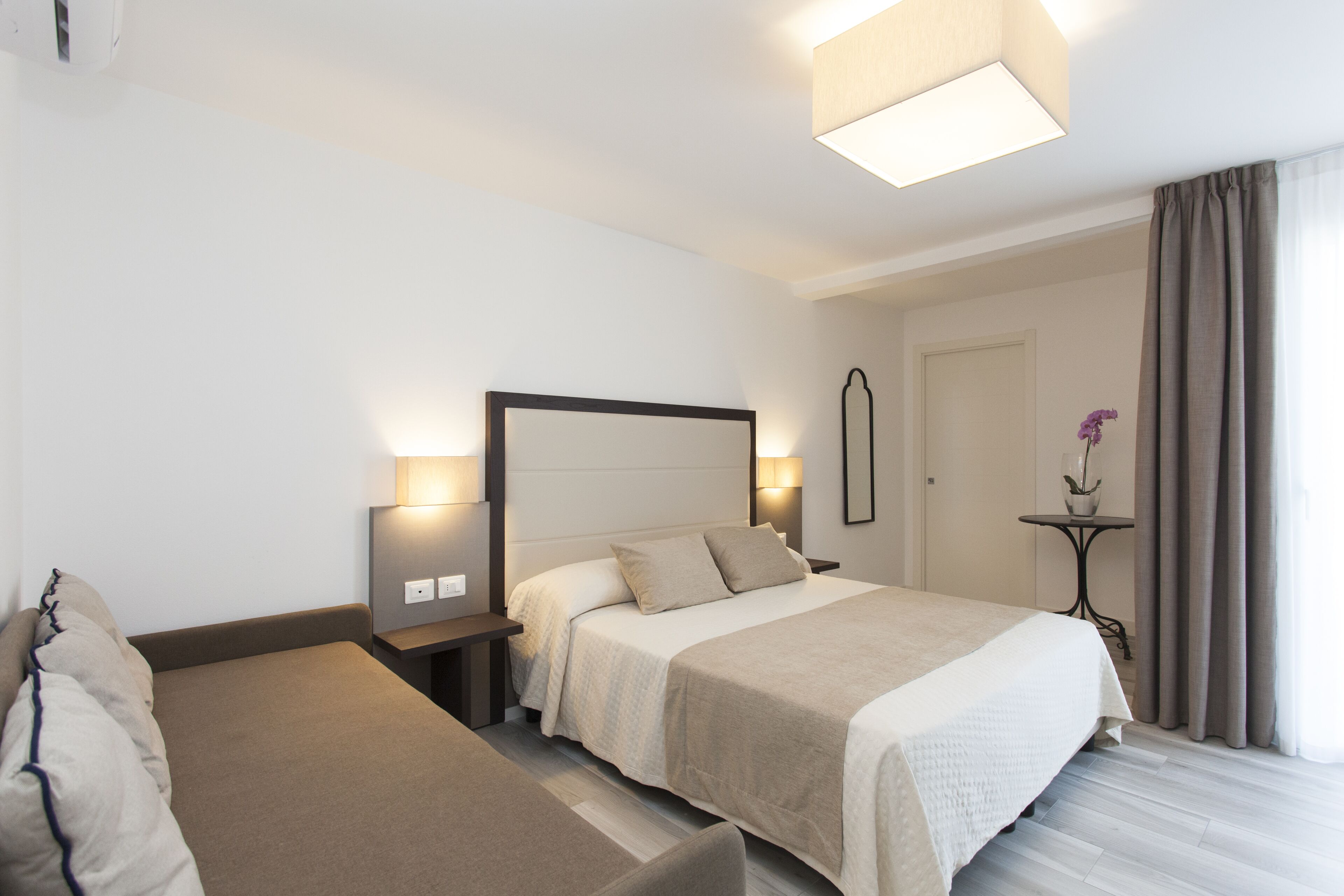 A hotel room with a bed, bedside tables, a nightstand, a lamp, a mirror, and a window with curtains.