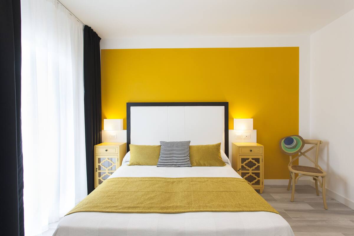 Habitación de hotel con cama, dos mesitas de noche, una silla y una pared decorada en amarillo.