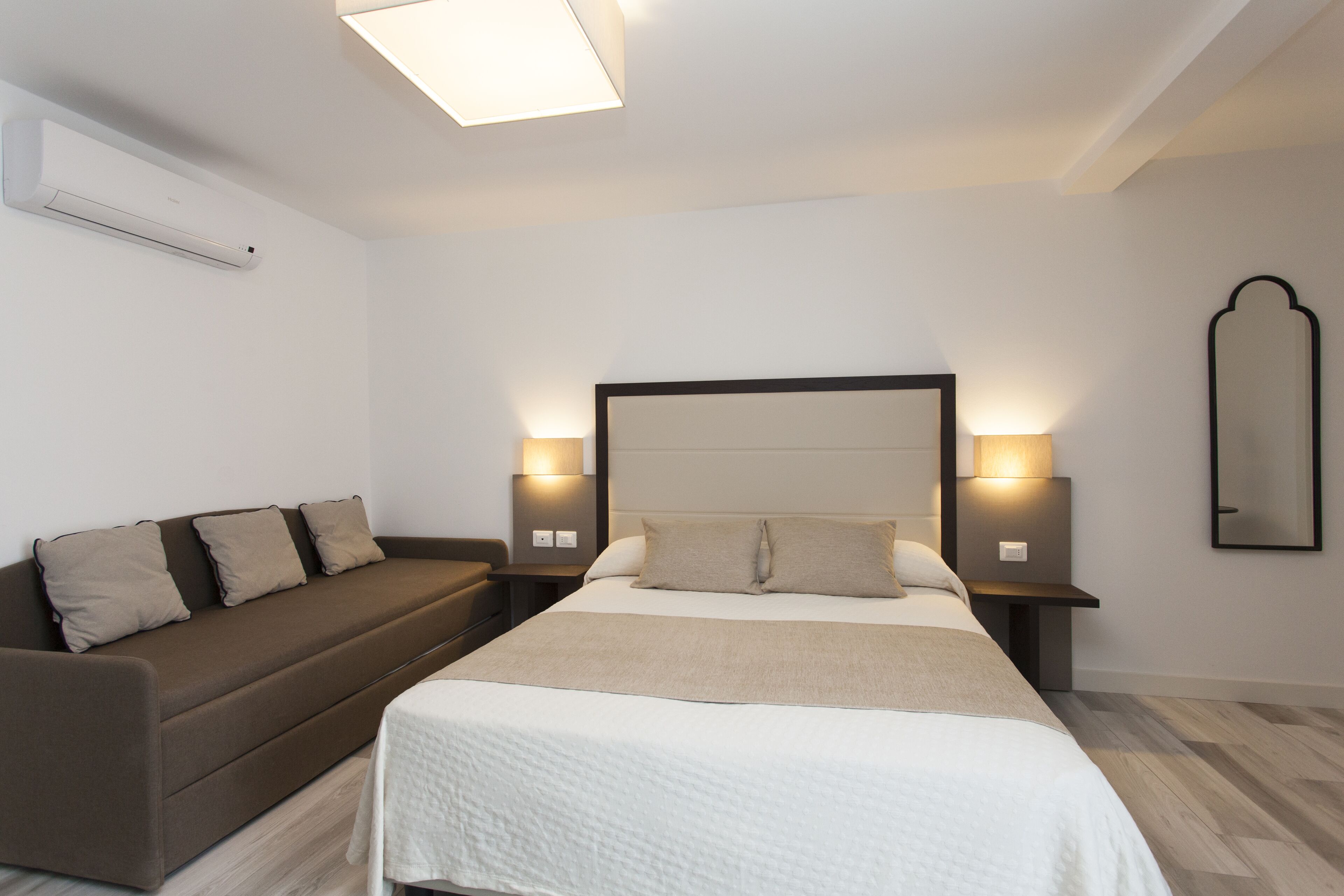 A modern hotel room with a bed, a sofa, and bedside tables with lamps.