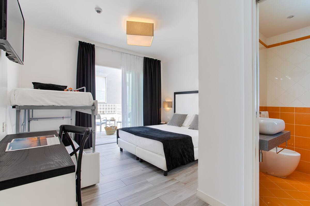 Una habitación de hotel con una cama individual superpuesta, un escritorio, una silla, una cama con colcha blanco y negro, una mesita de noche con lámpara y un baño con pared de azulejos naranja.