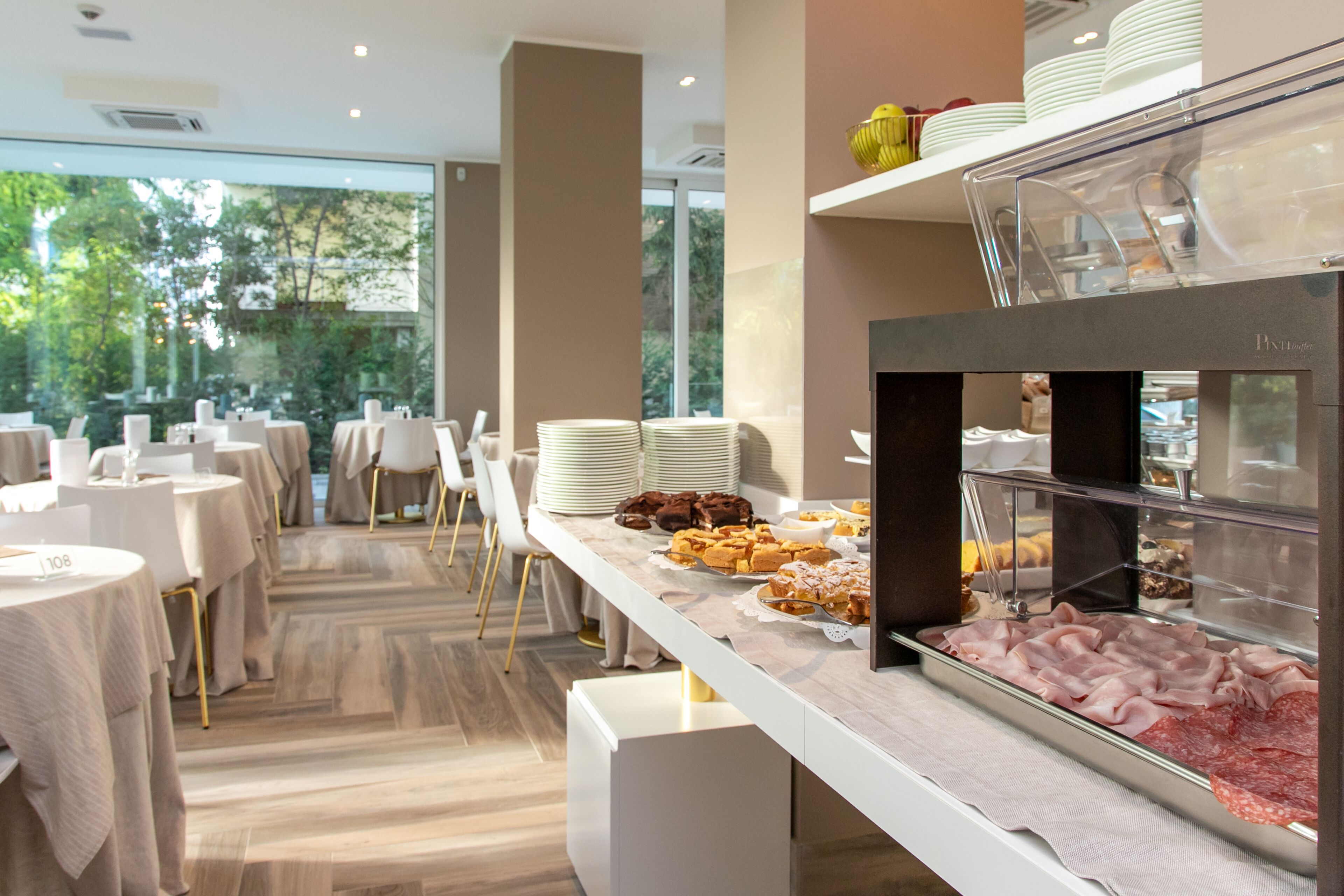 Zona de buffet amb diversos plats, incloent carns i pastissos, en un ambient modern amb taules i cadires blanques.