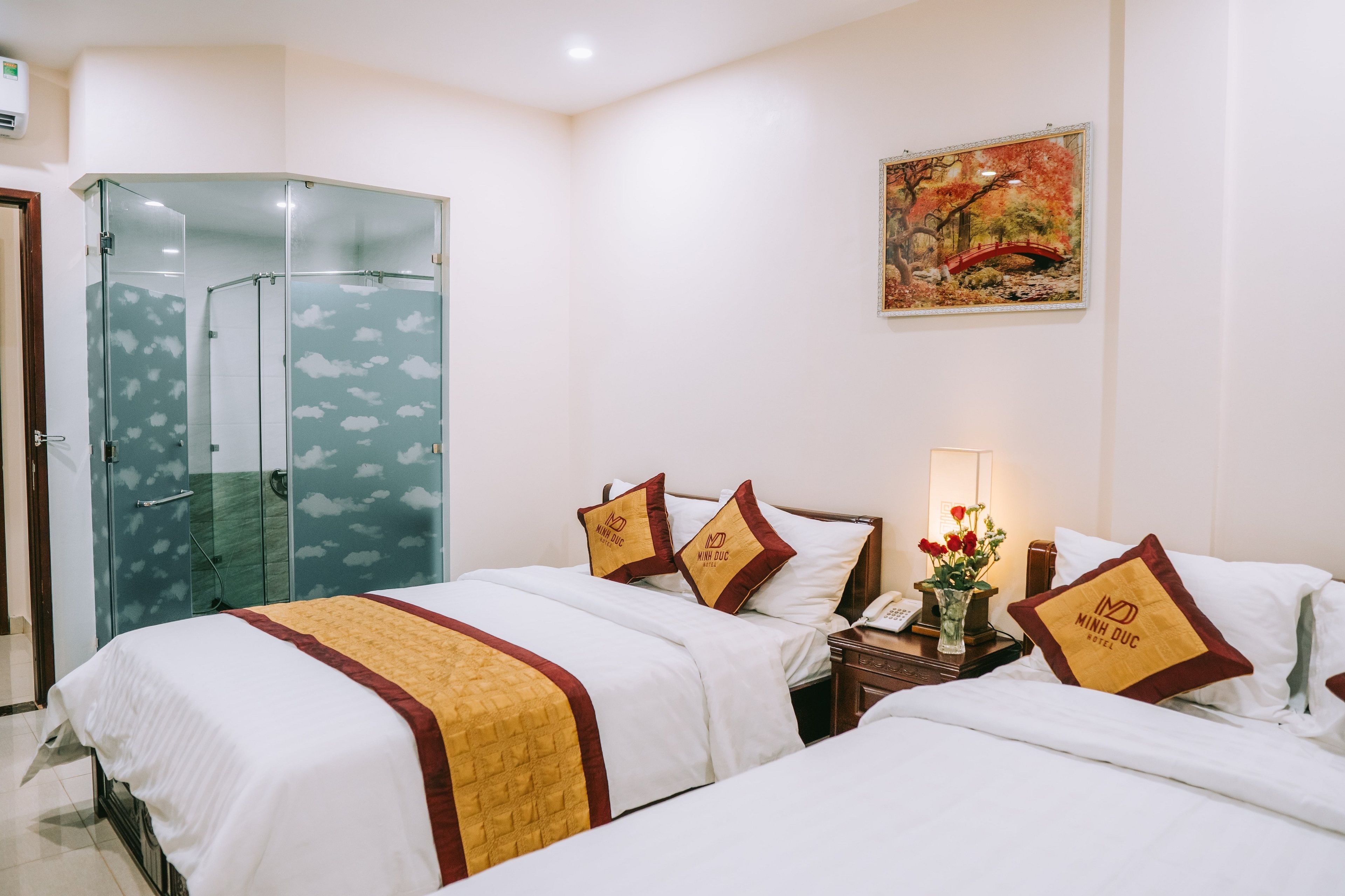 A hotel room with two beds, a glass shower enclosure, a nightstand with a lamp and phone, and a painting on the wall.
