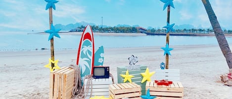 Montaje en la playa con tablas de surf, un televisor y cajas de madera, probablemente para un complejo turístico.