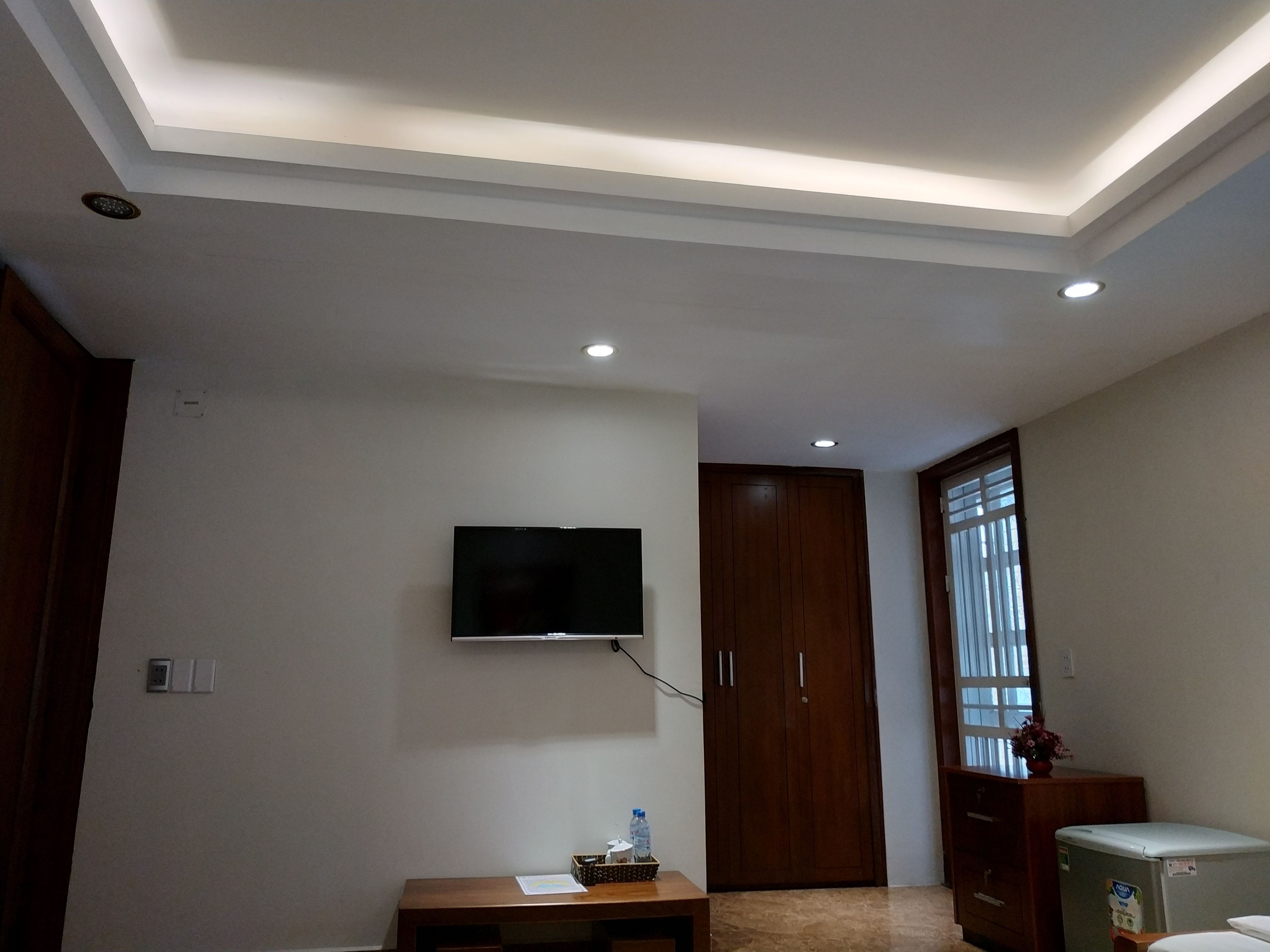 A room with a flat-screen TV mounted on the wall, a wooden wardrobe, a small table with a bottle, and a refrigerator.
