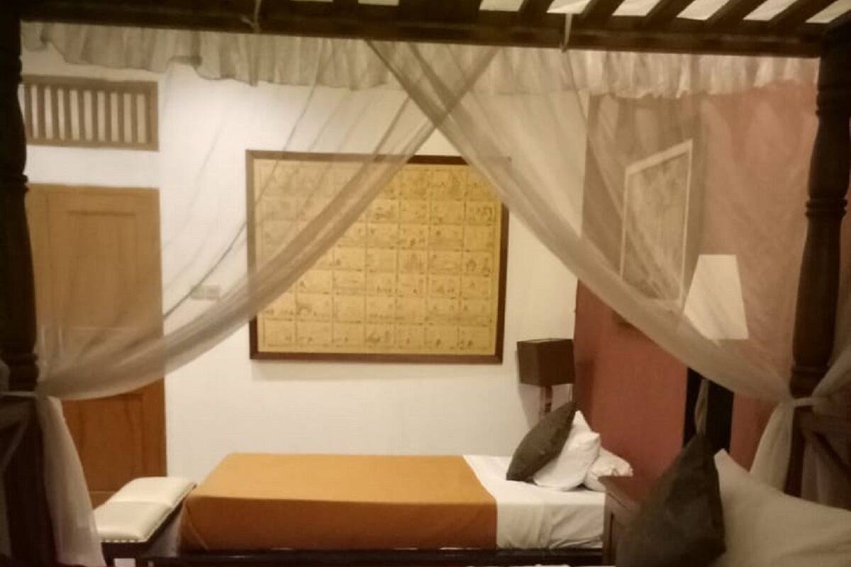 A room with a bed, a small table, and a chair. There is a window with a curtain and a wall-mounted lamp.