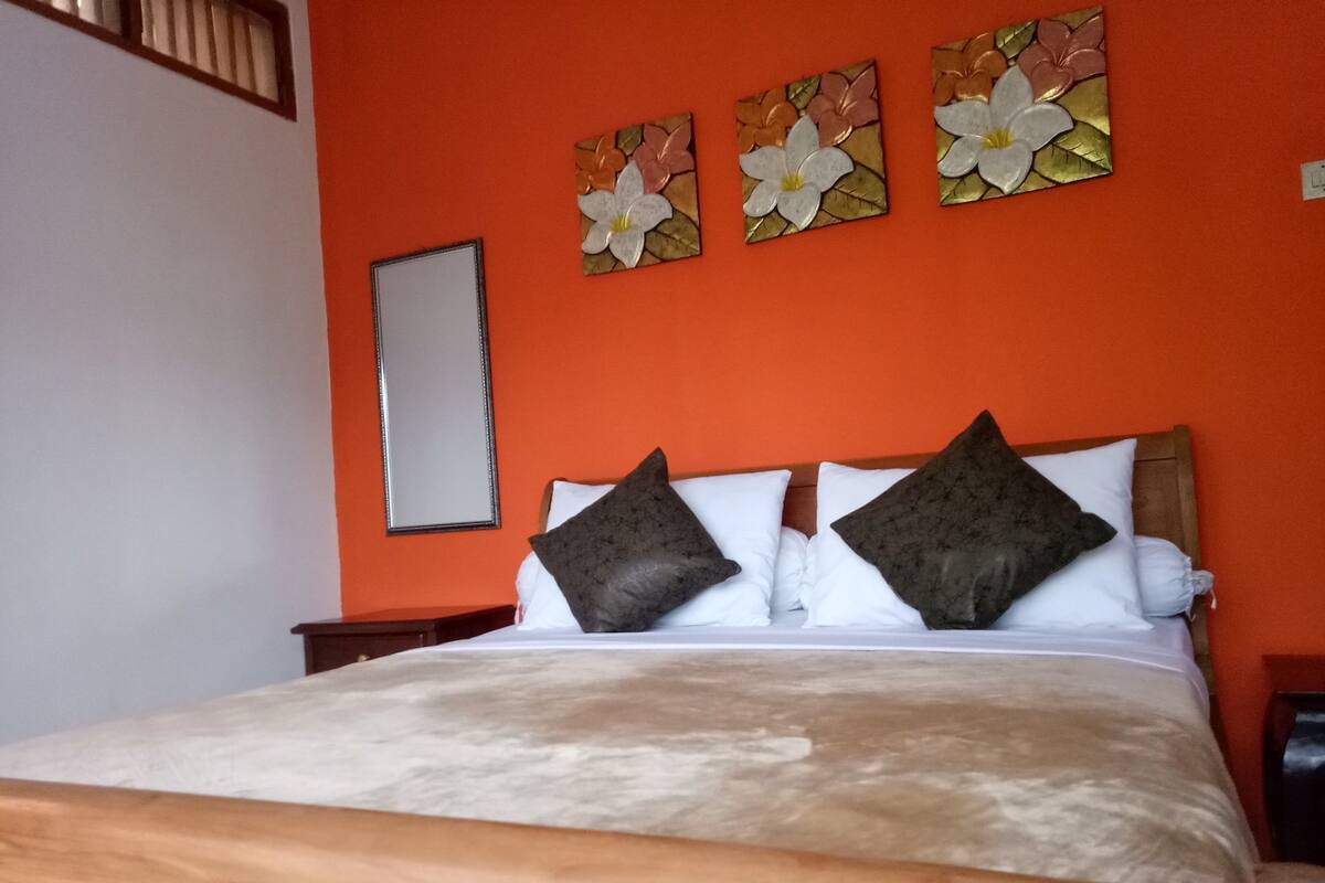 A bed with a wooden headboard, white bedding, and two dark pillows. There are three flower paintings on the orange wall above the bed.