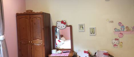 A room with a wooden wardrobe, a desk with a mirror, two Hello Kitty chairs, and a wall with Hello Kitty decorations.