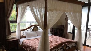 Una cama con dosel sobre soportes de madera y cortinas blancas, ubicada en una habitación con un ventanal y vistas a la vegetación.