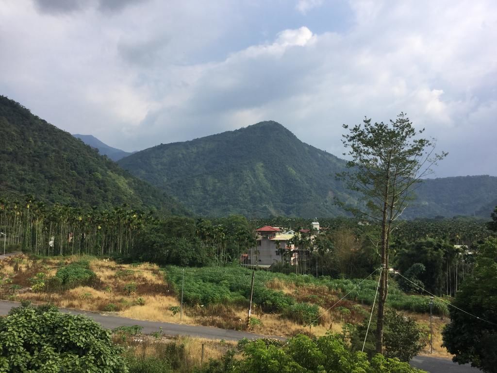 Paesaggio rurale con una strada, un edificio dal tetto rosso e dolci colline verdi sullo sfondo.